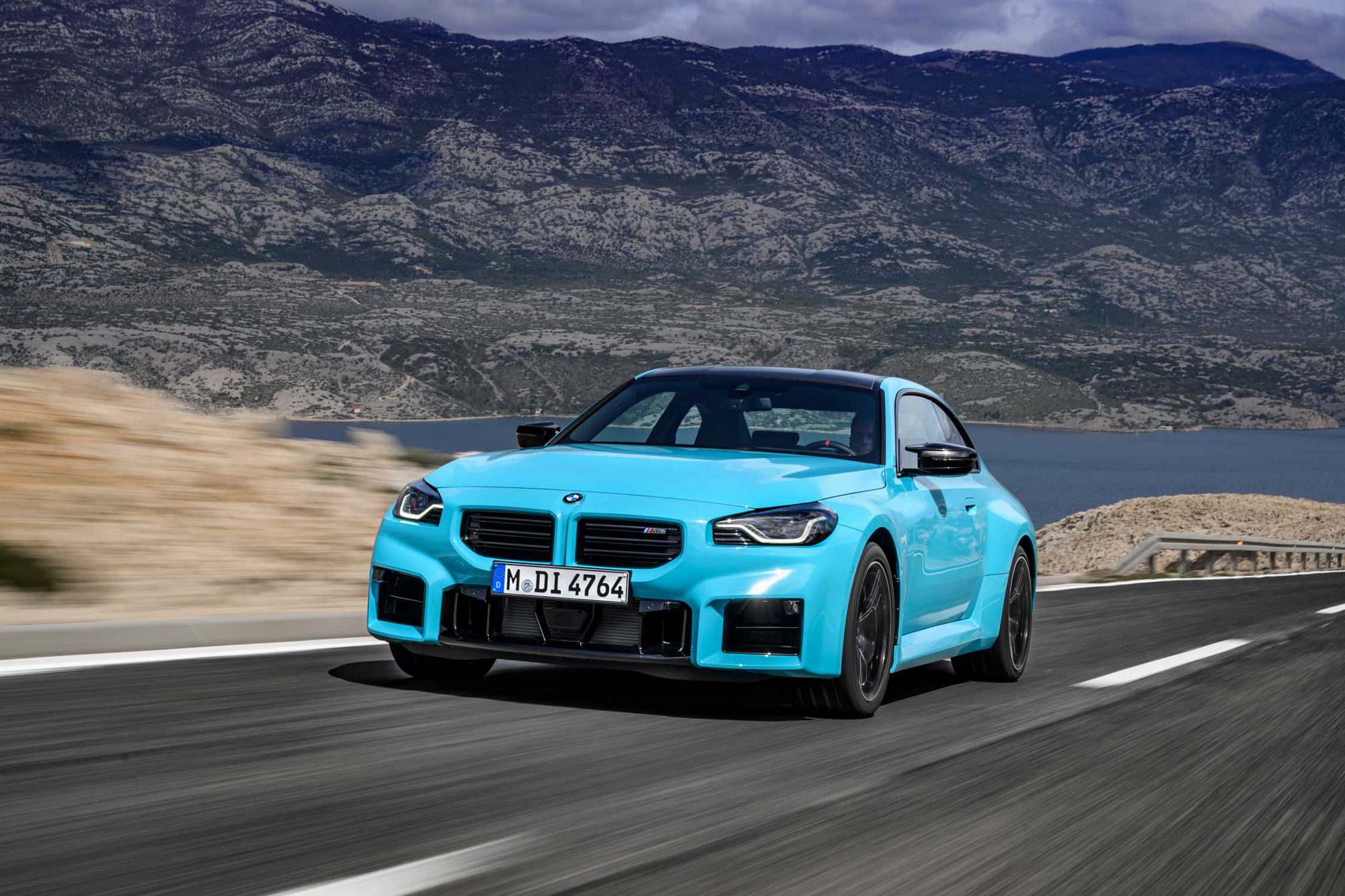 Τιρκουάζ μπλε BMW M2 σε πλάγια όψη από εμπρός αριστερά, σε δρόμο πάνω από θαλάσσιο κόλπο