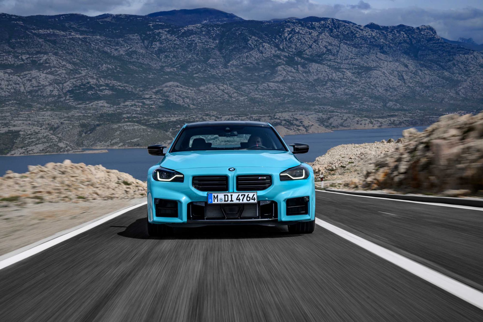 Τιρκουάζ μπλε BMW M2 κινείται μετωπικά σε έναν δρόμο, πίσω του μια γυμνή οροσειρά και θάλασσα